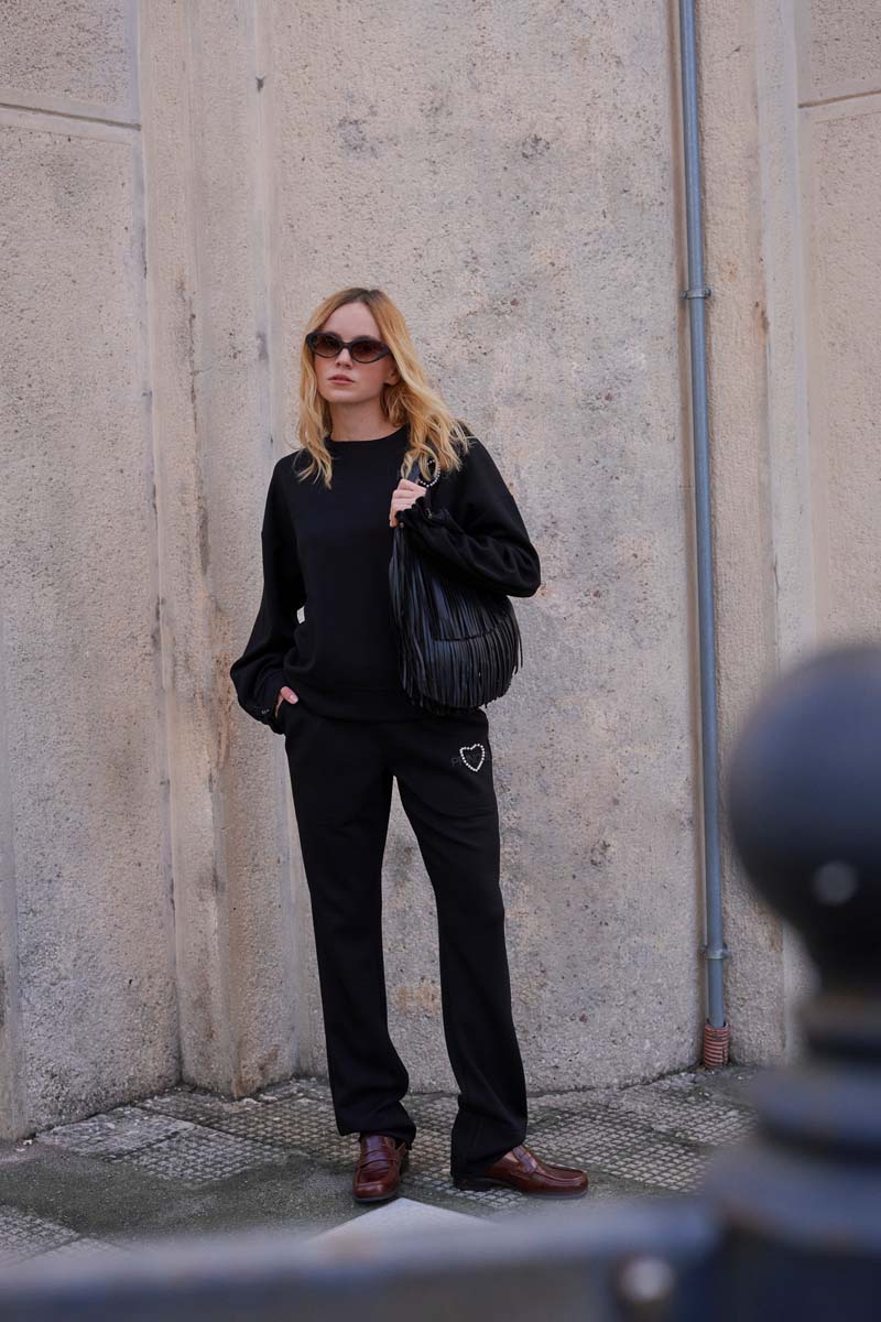 Eine Frau mit schwarzer Sonnenbrille lehnt an einer beigen Wand. Sie trägt ein schwarzes Sweatshirt und eine Sweathose von Princess Goes Hollywood mit einem elastischen Bund und einem silbernen Herz, dazu braune Schuhe und eine große gefaltete Handtasche.