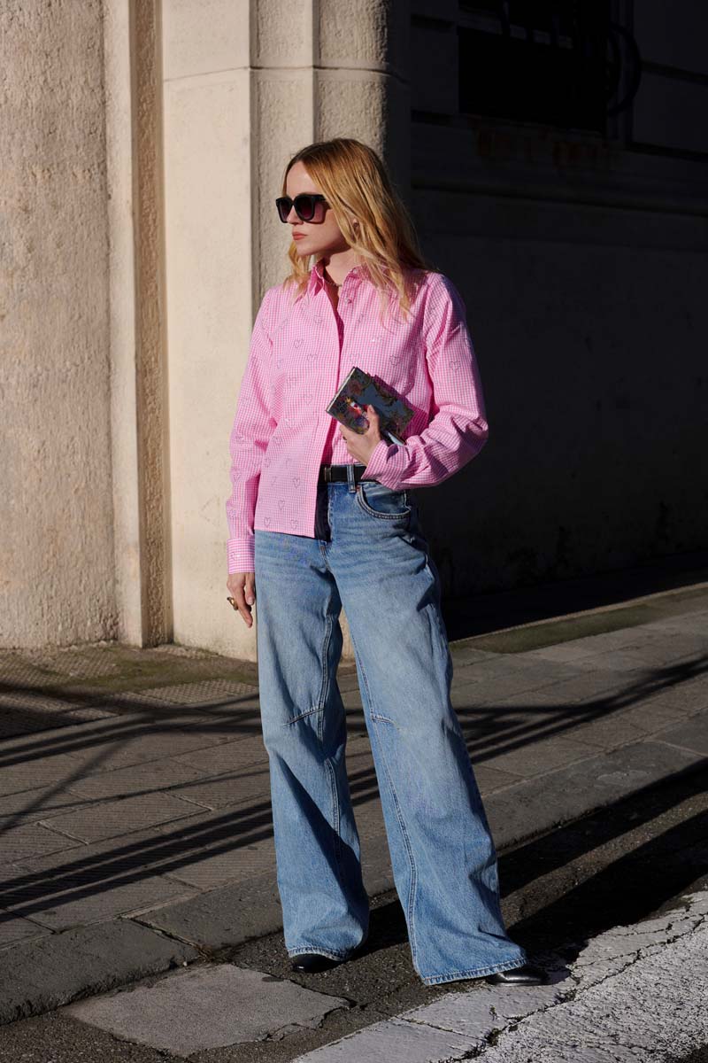 Eine Frau mit Sonnenbrille, einer hellrosa Princess Goes Hollywood Baumwolle Hemdbluse und weit geschnittenen Blue Jeans steht an einem sonnigen Tag draußen und hält eine gemusterte Clutch. Ihr langes blondes Haar fängt das Sonnenlicht auf dem Bürgersteig ein.