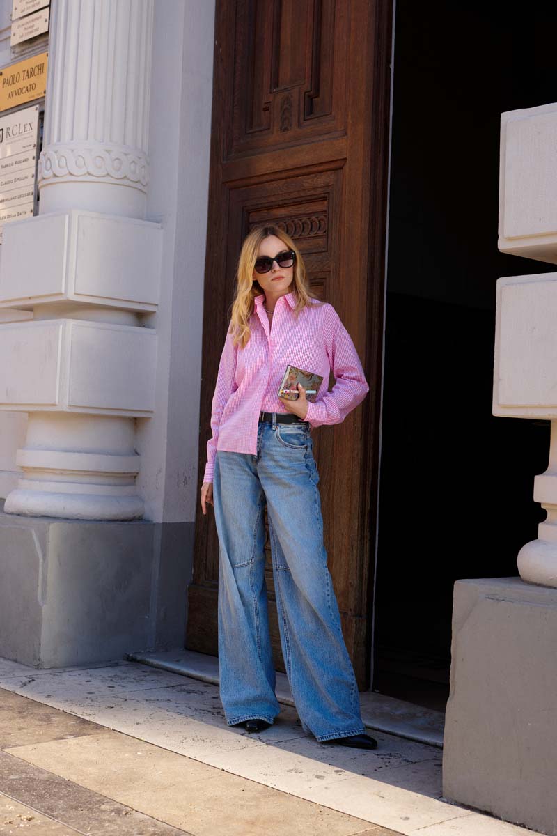 Eine Frau steht in einer Tür und trägt eine leuchtend pinke Princess Goes Hollywood Baumwolle Hemdbluse und eine weit geschnittene Bluejeans, dazu eine kleine Clutch und eine Sonnenbrille. Weiße Säulen und eine große Holztür vervollständigen den klassischen architektonischen Hintergrund.