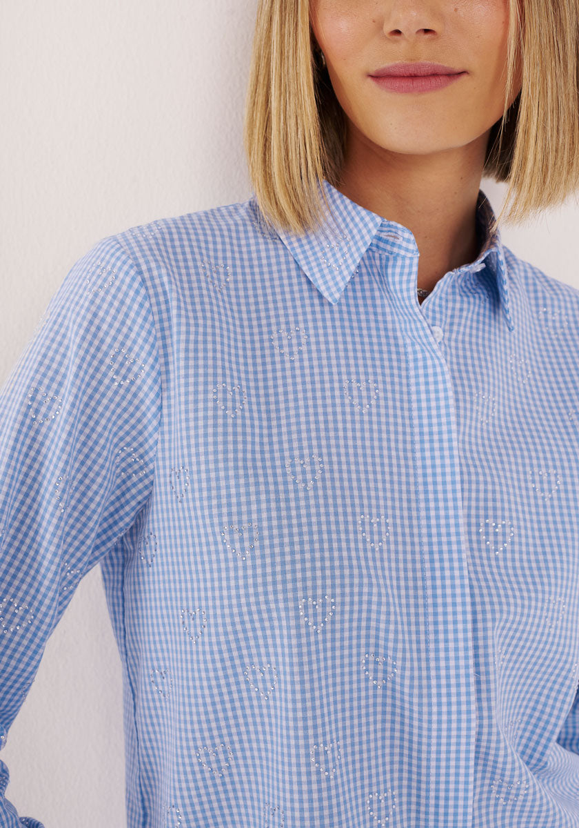 Eine Frau mit blondem Bubikopf trägt eine hellblau-weiß karierte Bluse von Princess Goes Hollywood, die mit dezenten Strassherzmotiven verziert ist. Nur ihr unteres Gesicht, ihr Hals und ihr oberer Oberkörper sind zu sehen, während sie leicht lächelt.