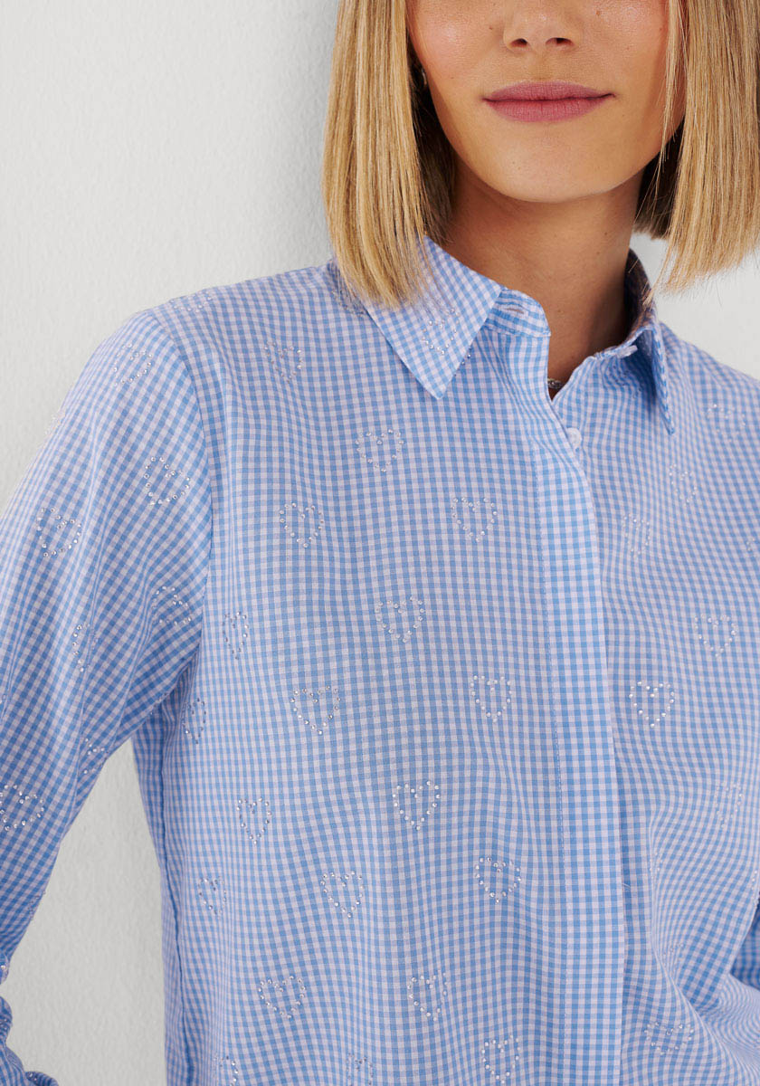 Eine Frau mit glattem blondem Haar trägt eine hellblau-weiße Vichy-Karo-Bluse mit kleiner Blumenstickerei von Princess Goes Hollywood. An der Nase und in der Mitte des Oberkörpers abgeschnitten, lächelt sie sanft gegen eine blasse Wand.