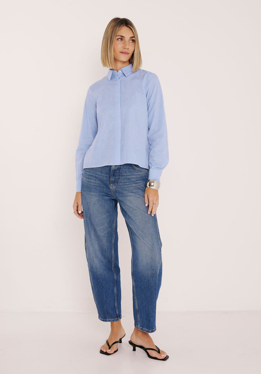 Eine Frau mit blondem Bubikopf steht vor einer weißen Wand. Sie trägt eine hellblau gestreifte Baumwollbluse von Princess Goes Hollywood, lässige Bluejeans, schwarze hochhackige Sandalen und ein silbernes Manschettenarmband. Sie blickt mit einem neutralen Blick nach vorne.