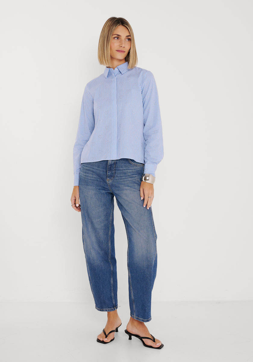 Eine Frau mit glattem, schulterlangem blondem Haar und neutralem Gesichtsausdruck steht vor einer weißen Wand und trägt eine hellblau gestreifte Baumwollbluse von Princess Goes Hollywood, lässige Bluejeans, schwarze hochhackige Sandalen und ein silbernes Armband.