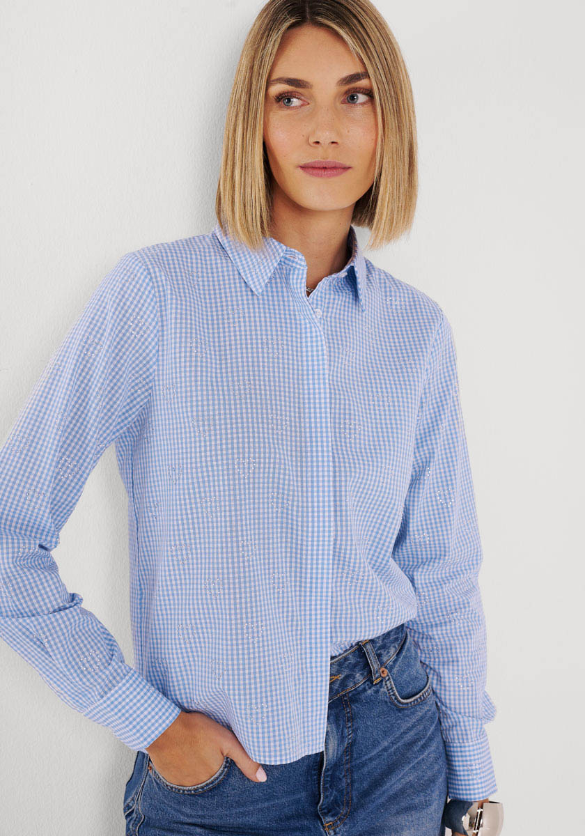 Eine Frau mit glattem, schulterlangem blondem Haar steht vor einer weißen Wand und trägt eine hellblau-weiß karierte Bluse von Princess Goes Hollywood, die in eine blaue Jeans gesteckt ist. Eine Hand steckt in ihrer Hosentasche, während sie mit neutralem Gesichtsausdruck leicht nach rechts schaut.