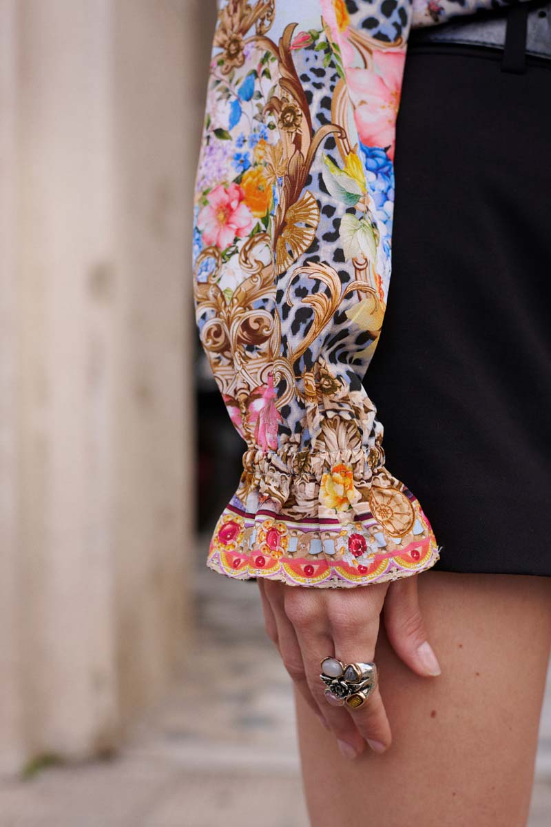 Eine Nahaufnahme eines Arms zeigt eine farbenfrohe Princess Goes Hollywood Seidenbluse mit floralem Print und barocken Mustern, die in einer gerüschten Manschette endet. Die Person trägt dazu schwarze Shorts und einen großen, verschlungenen Ring mit Schmucksteinen.