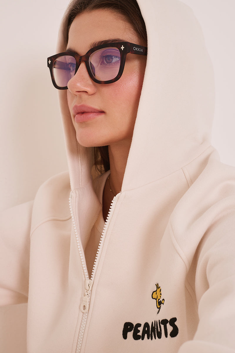 Eine Frau mit langen braunen Haaren trägt eine modische Okkia-Lesebrille mit quadratischem Gestell und dunklen Rändern sowie ein cremefarbenes Sweatshirt mit Kapuze und Reißverschluss mit einem kleinen gelben Woodstock-Cartoon und dem Text "PEANUTS". Sie blickt nach links, Kapuze hochgezogen.