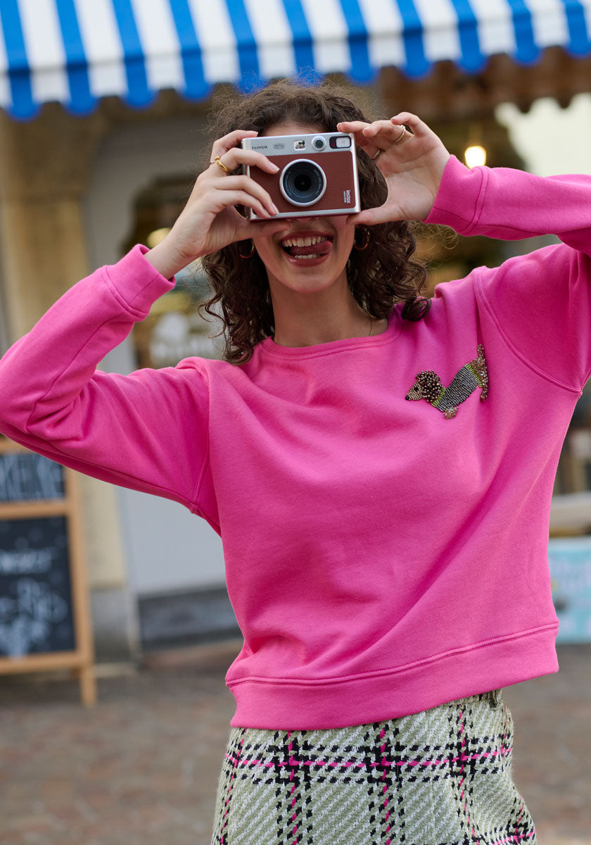 A smiling woman with curly hair wears a bright pink Princess Goes Hollywood Sweatshirt and a patterned skirt with Perlenstickerei, holding a camera up to her face under a blue-and-white striped awning outdoors.