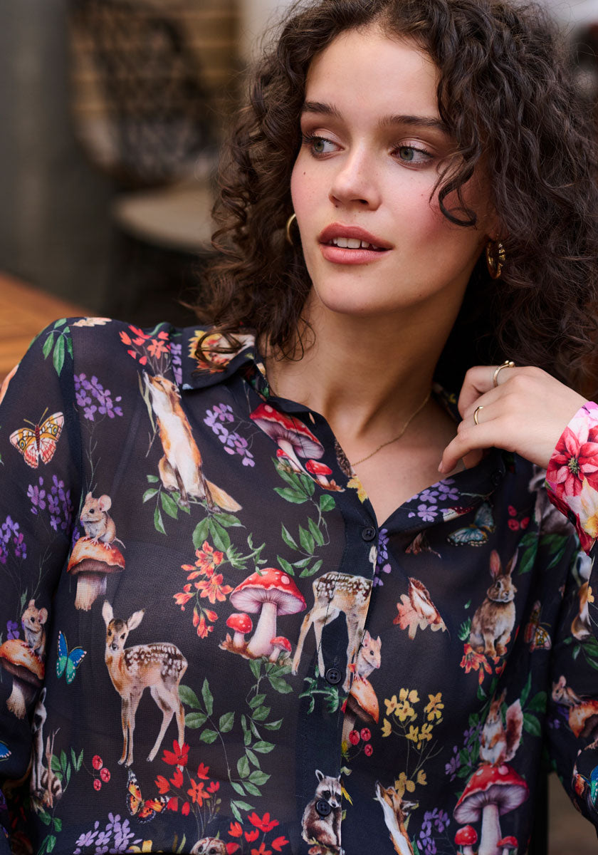 A young woman with curly brown hair and gold hoops looks to the side, wearing a whimsical Waldtier-Print Hemdbluse by Princess Goes Hollywood featuring deer, mushrooms, and butterflies. A softly blurred background highlights her stylish shirt.