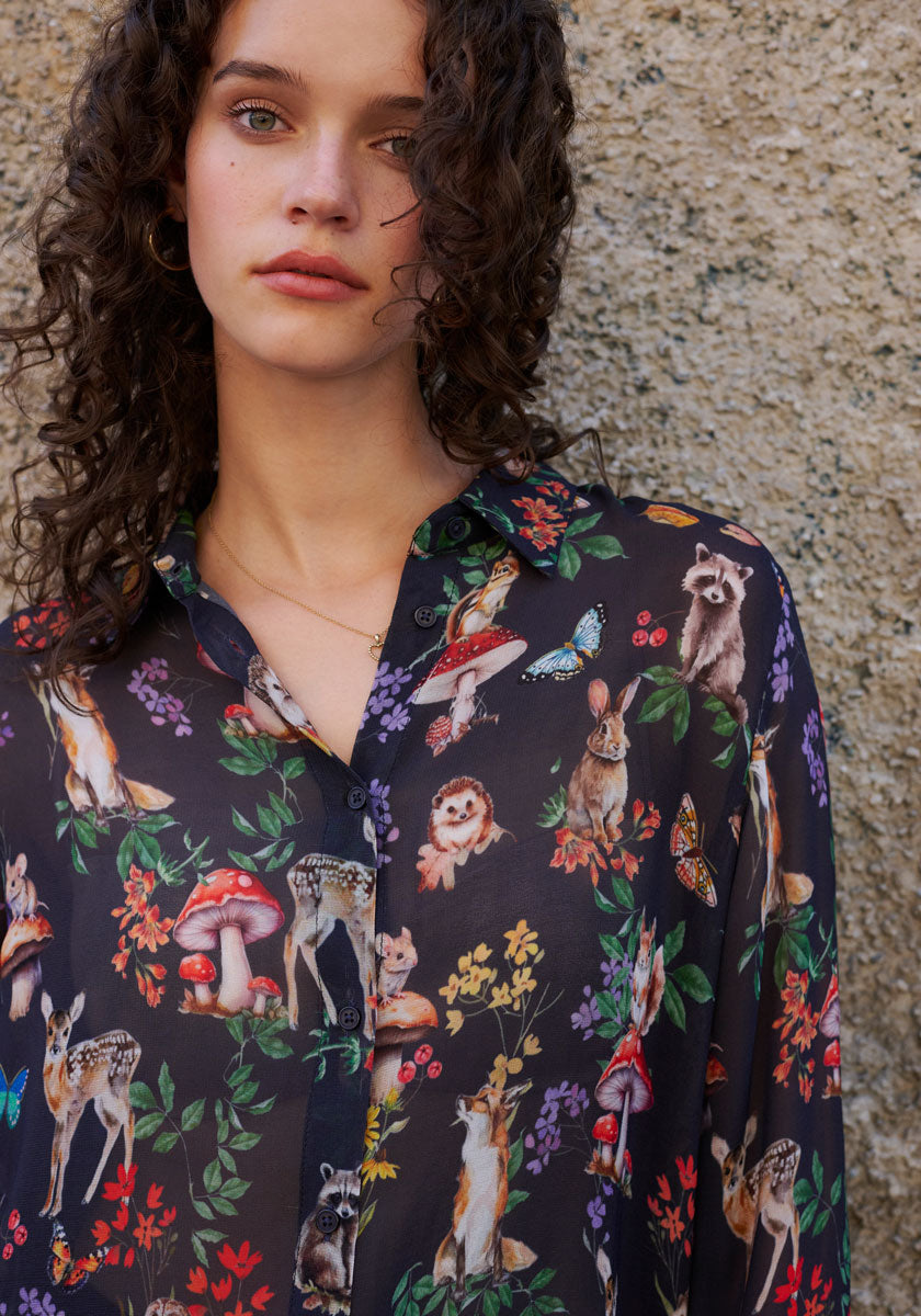 A young woman with curly brown hair stands by a textured stone wall, wearing a dark Princess Goes Hollywood Hemdbluse adorned with colorful woodland animals, mushrooms, flowers, and butterflies in a charming Waldtier-Print.