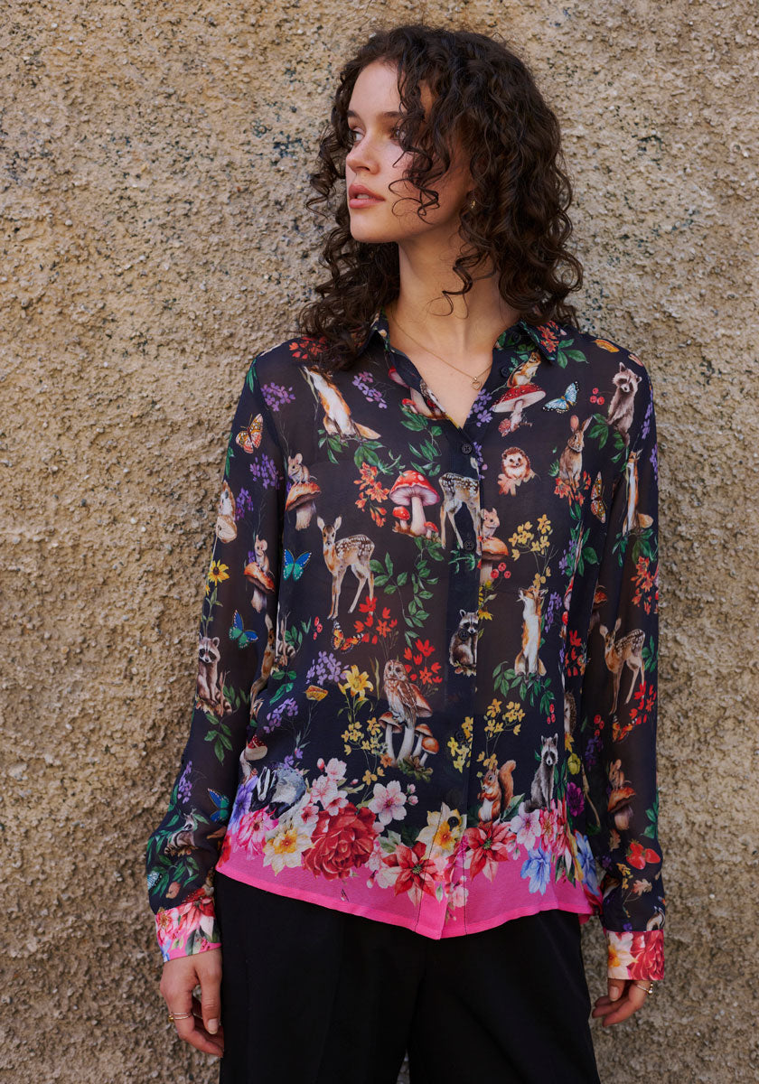 A woman with curly hair stands by a textured beige wall wearing a vibrant Princess Goes Hollywood Waldtier-Print Hemdbluse with colorful flowers, animals, mushrooms, and butterflies. The blouse features a pink floral hem as she gazes thoughtfully to her left.