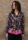 A woman with curly hair stands by a textured beige wall wearing a vibrant Princess Goes Hollywood Waldtier-Print Hemdbluse with colorful flowers, animals, mushrooms, and butterflies. The blouse features a pink floral hem as she gazes thoughtfully to her left.