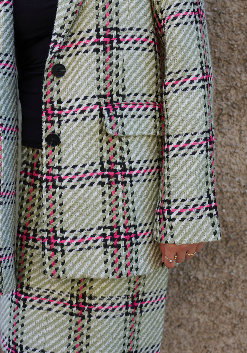 Wearing a Princess Goes Hollywood textured Tweed Blazer in light green, black, white, and pink Karo-Print with black buttons and a pocket over a black shirt, gold rings gleam on their hand by a rugged stone wall.