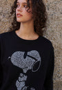 A young woman with curly brown hair stands by a stone wall, wearing a black Princess Goes Hollywood Snoopy Sweatshirt aus Baumwolle with a white strassverzierung graphic. Soft natural light highlights her upper body as she gazes to the side.