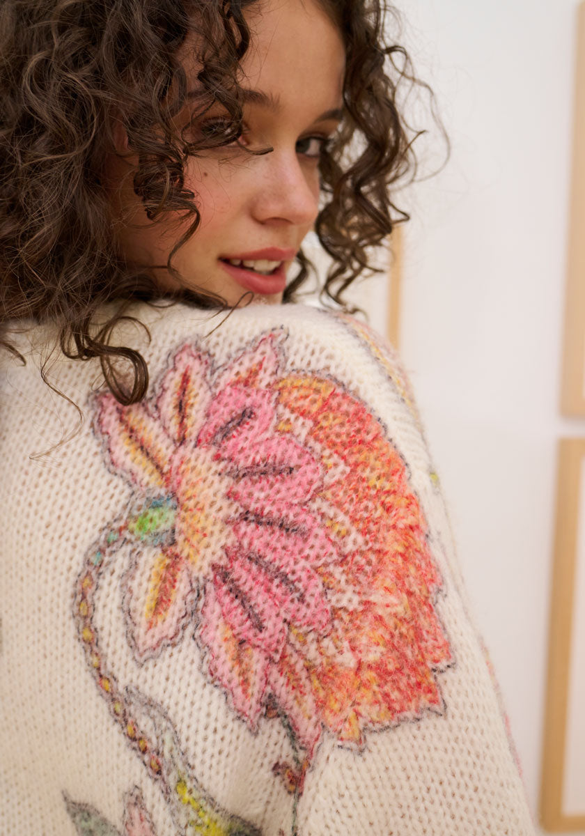 A young woman with curly brown hair smiles over her shoulder, wearing a cream Princess Goes Hollywood Wolle-Alpaka-Mix Pullover adorned with a large pink and orange flower on the back. She stands before softly lit, light-colored panels.