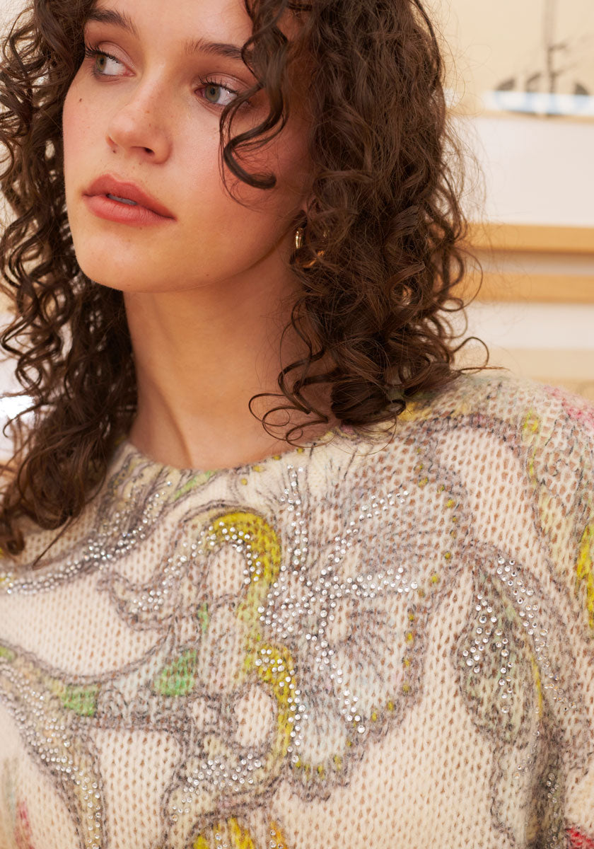 A young woman with curly brown hair wears a Princess Goes Hollywood Wolle-Alpaka-Mix Pullover adorned with sparkling Strassverzierung. She gazes calmly to the side, soft natural light illuminating her clear skin and gentle features.