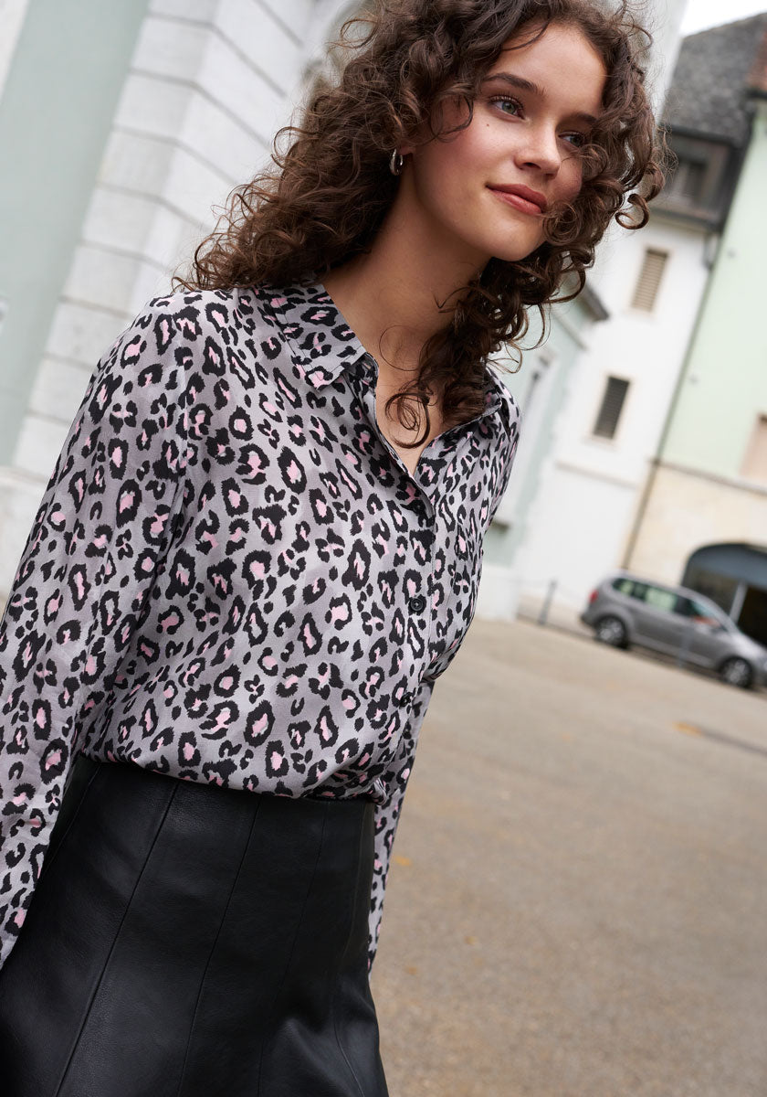 A woman with curly brown hair smiles outdoors, wearing a Frogbox by Princess Leo-Print Bluse with Hemdkragen tucked into a high-waisted black leather skirt. She stands in an urban setting with pale buildings and a parked car behind her.