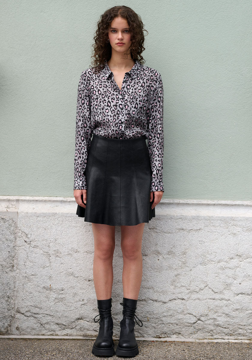 A young woman with curly hair stands against a pale green wall, wearing a Frogbox by Princess Leo-Print Bluse with Hemdkragen tucked into a black leather A-line skirt and black lace-up boots. Her expression is neutral, arms at her sides.