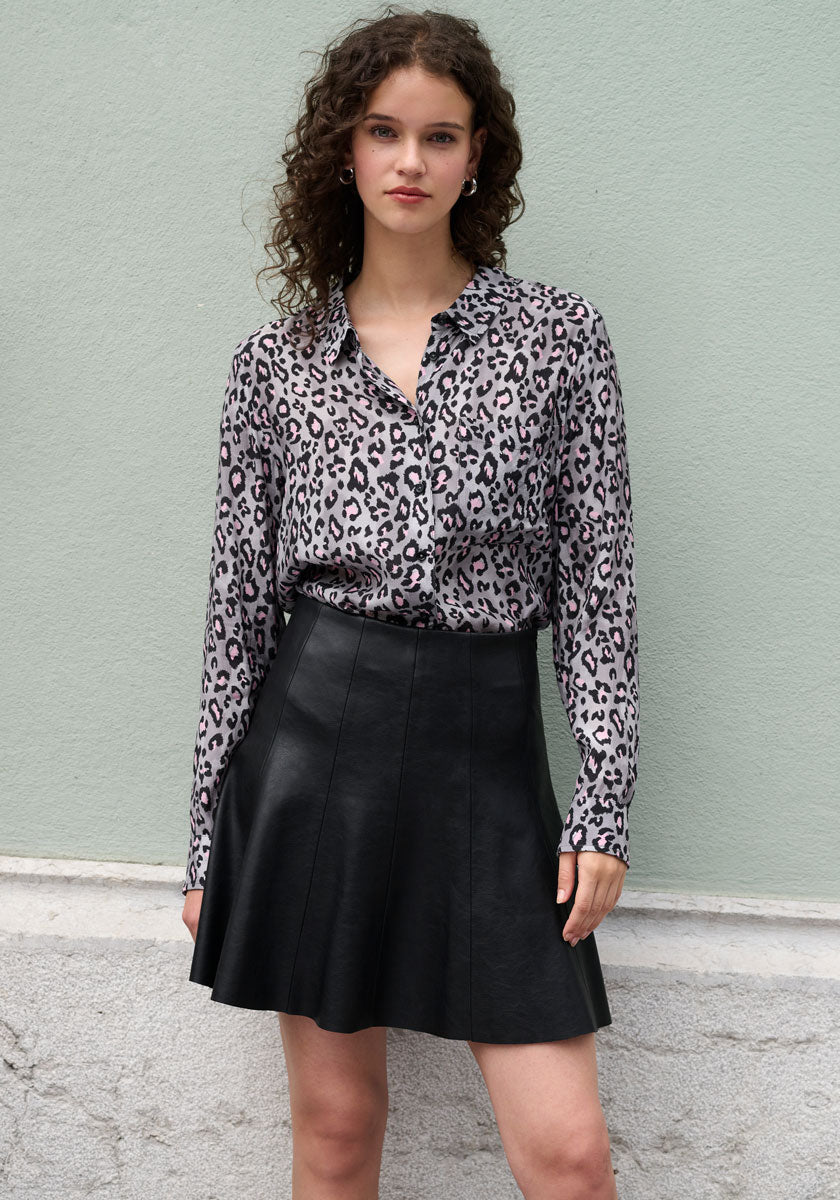 A young woman with curly brown hair stands against a pale green wall, wearing a Frogbox by Princess Bluse with Leo-Print and Hemdkragen, tucked into a high-waisted black flared leather skirt. She gazes at the camera with a neutral expression.