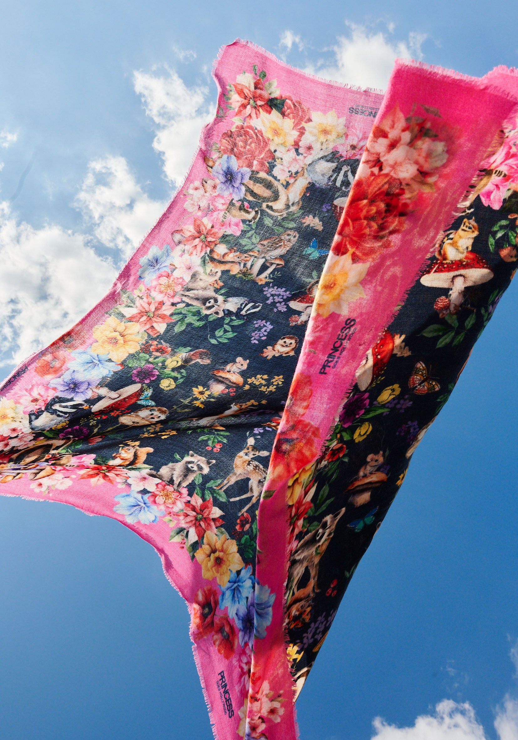 A colorful quadratischer Schal by Princess Goes Hollywood, featuring pink edges and multicolored floral patterns, billows against a blue sky as sunlight shines through its fabric.