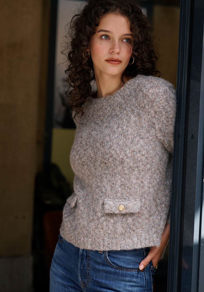 A woman with curly brown hair and gold hoop earrings stands by a doorway in blue jeans, wearing a beige Princess Goes Hollywood Grobstrickpullover aus Bio-Baumwolle with two buttoned pockets. The softly blurred background is indoors.