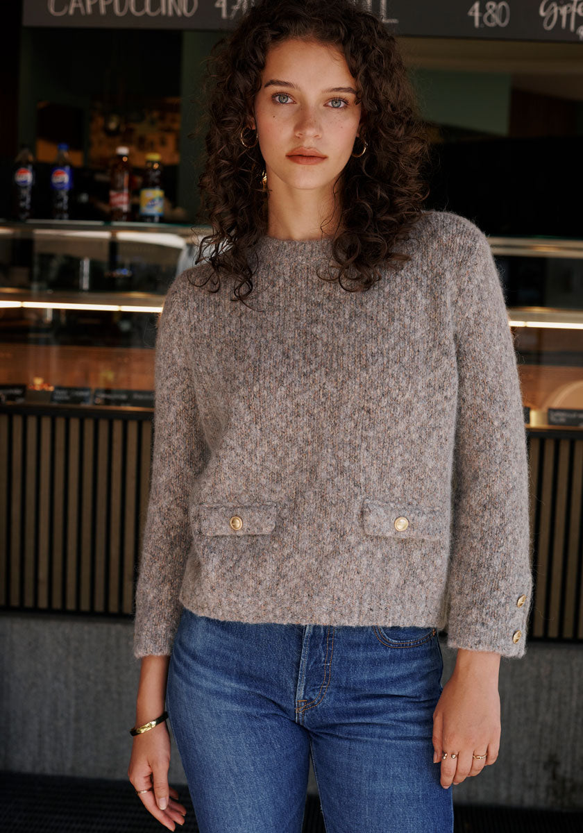 A woman with curly brown hair and blue eyes stands indoors, wearing a gray Princess Goes Hollywood Grobstrickpullover aus Bio-Baumwolle with gold buttons and blue jeans. She faces the camera; behind her are a cafe counter and blackboard menu.