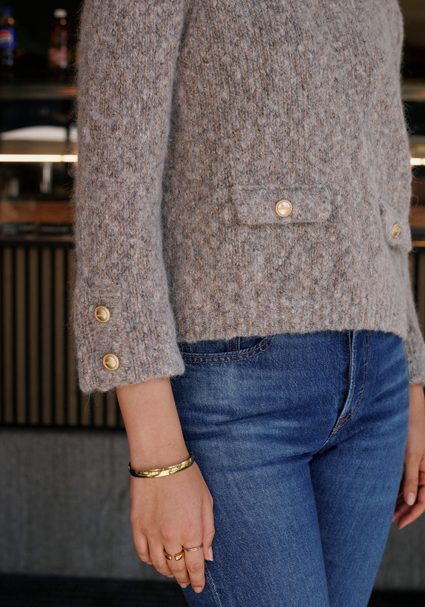 A person wears a light gray textured Grobstrickpullover by Princess Goes Hollywood, featuring gold buttons on the sleeves and pocket flaps, styled with blue jeans. They sport a gold bracelet and two rings. The indoor background appears blurred.