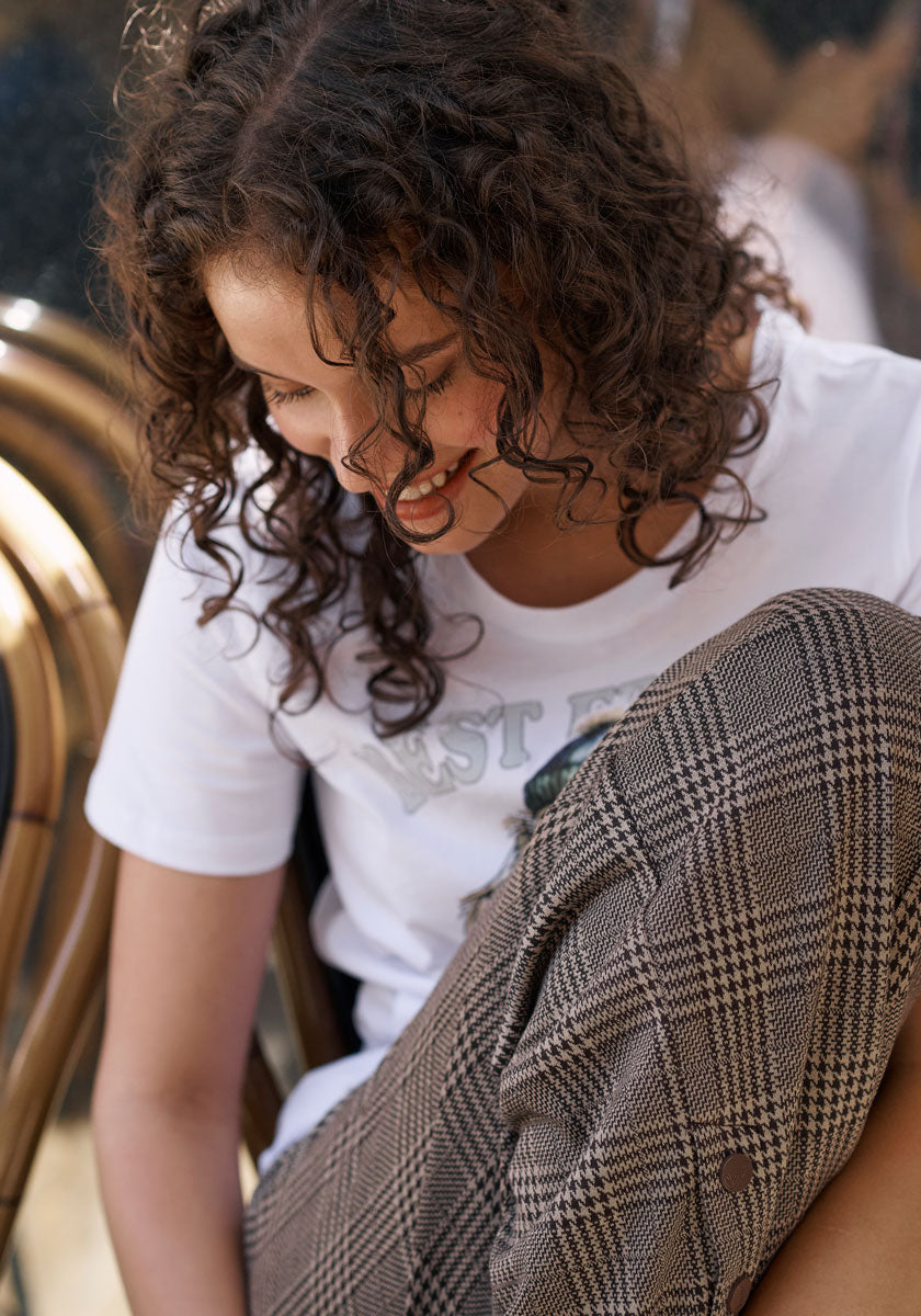 A young woman with curly hair smiles softly, wearing a white T-shirt and Princess Goes Hollywood checked trousers. Relaxed, she rests one arm on her raised knee amid stacked chairs and sunlight in the background.