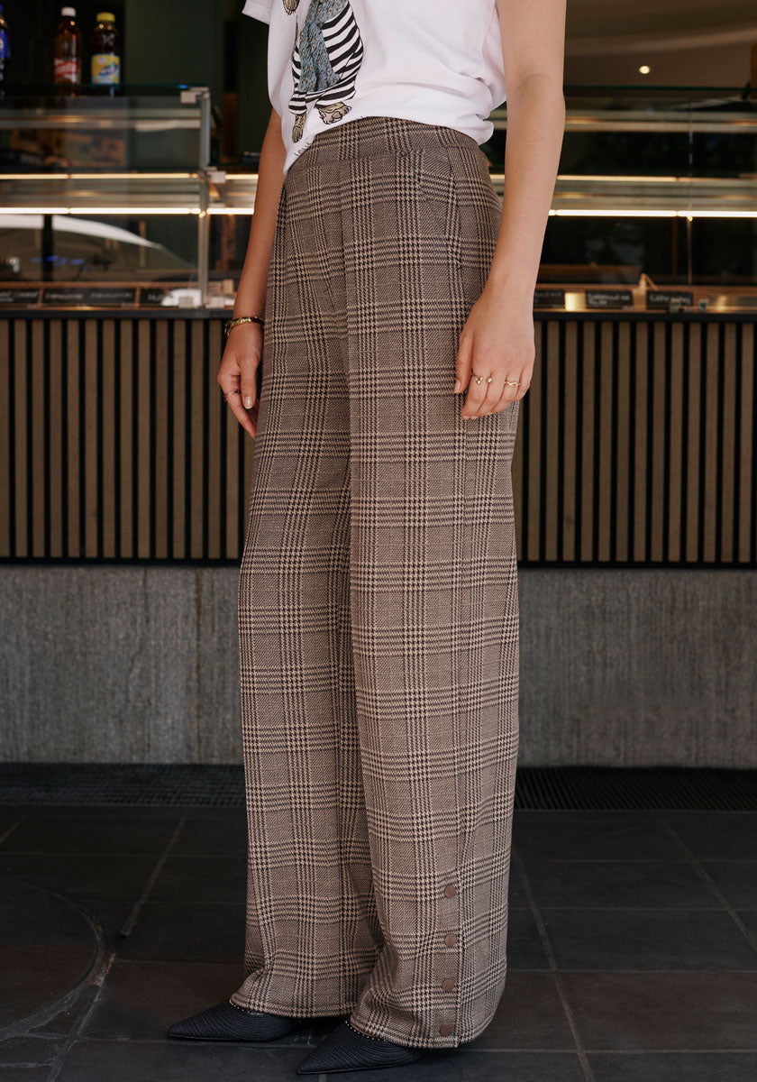 A person indoors wears high-waisted, wide-leg Princess Goes Hollywood trousers with brown and beige check pattern, paired with a white graphic tee. Only the body from shoulders down is shown against a modern café counter backdrop.