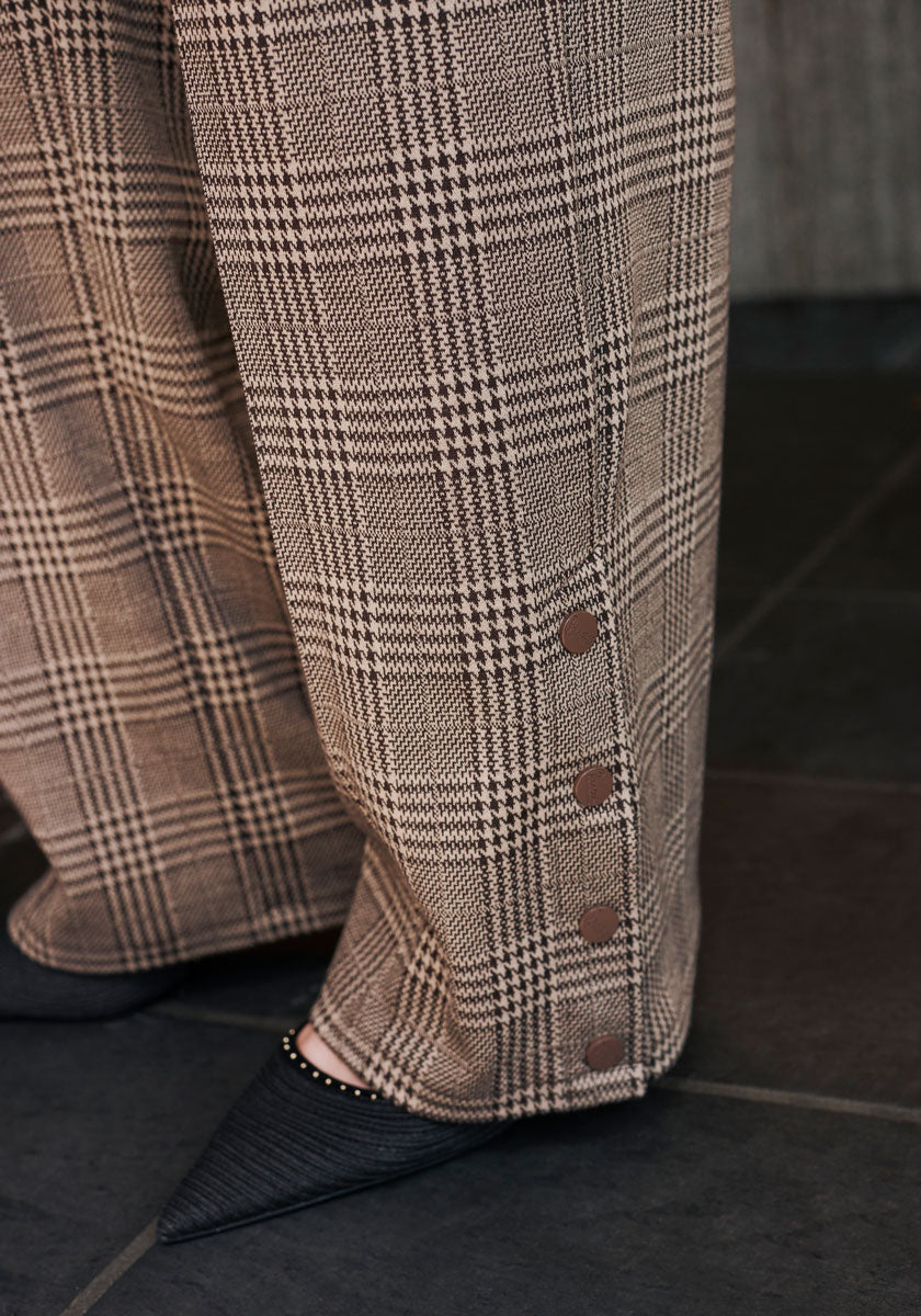 A close-up of brown and beige checked wide-leg pants by Princess Goes Hollywood with four round buttons at the ankle, styled with black pointed-toe studded shoes on a dark tiled floor.