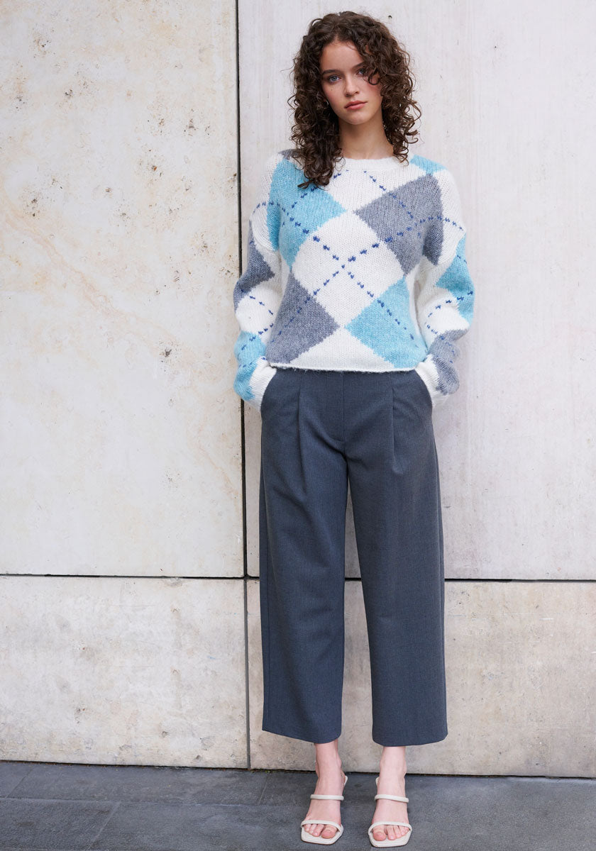 A woman with curly brown hair stands against a light stone wall, wearing a fluffy Princess Goes Hollywood Strickpullover aus Wolle-Alpaka-Mix with blue, gray, and white argyle pattern, loose gray cropped trousers, and white open-toe sandals. She gazes forward calmly.