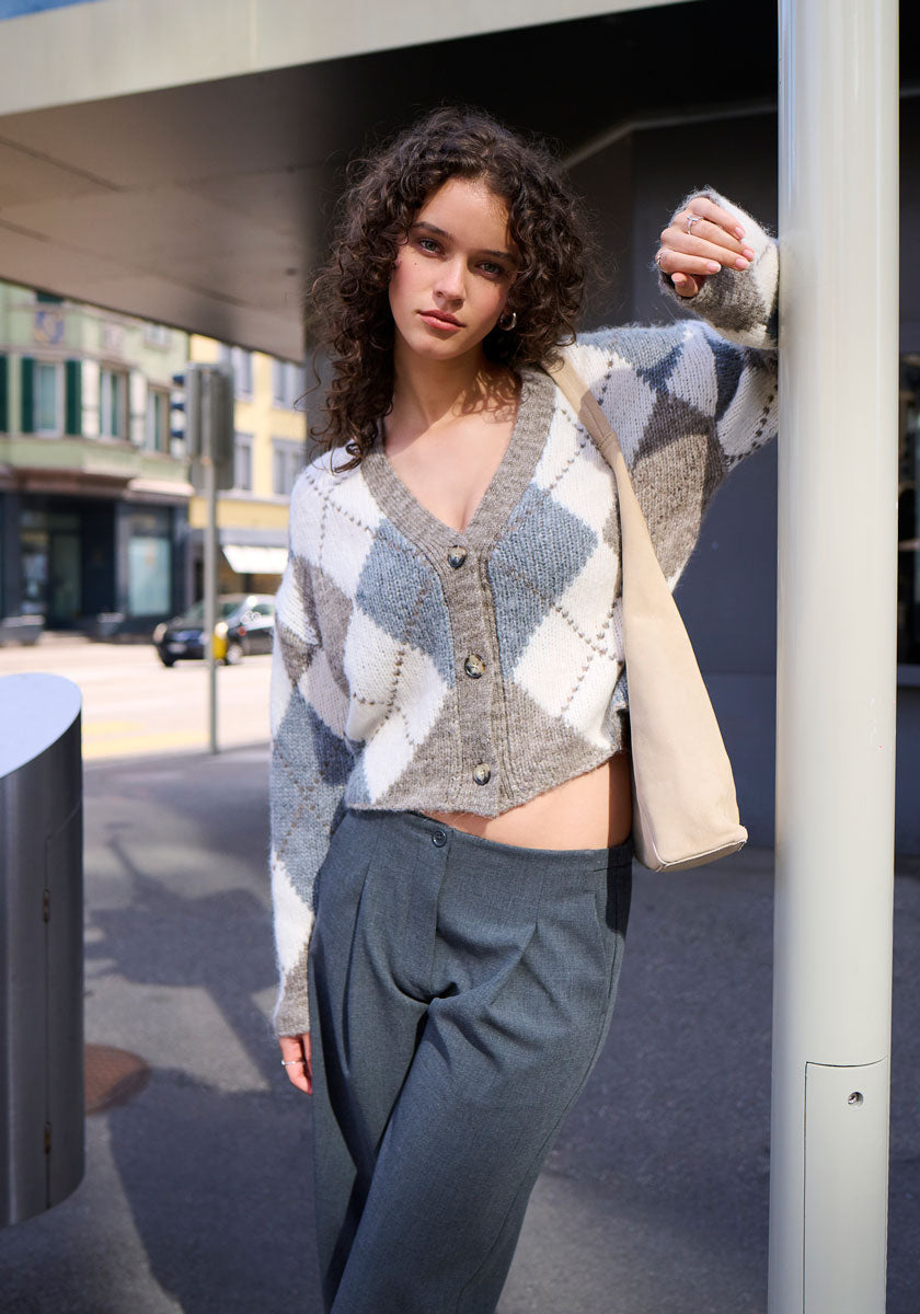 A young woman with curly brown hair leans against a city pole, wearing a Princess Goes Hollywood grey-white oversized argyle cardigan, blue-grey 7/8-length trousers, and a cream shoulder bag.