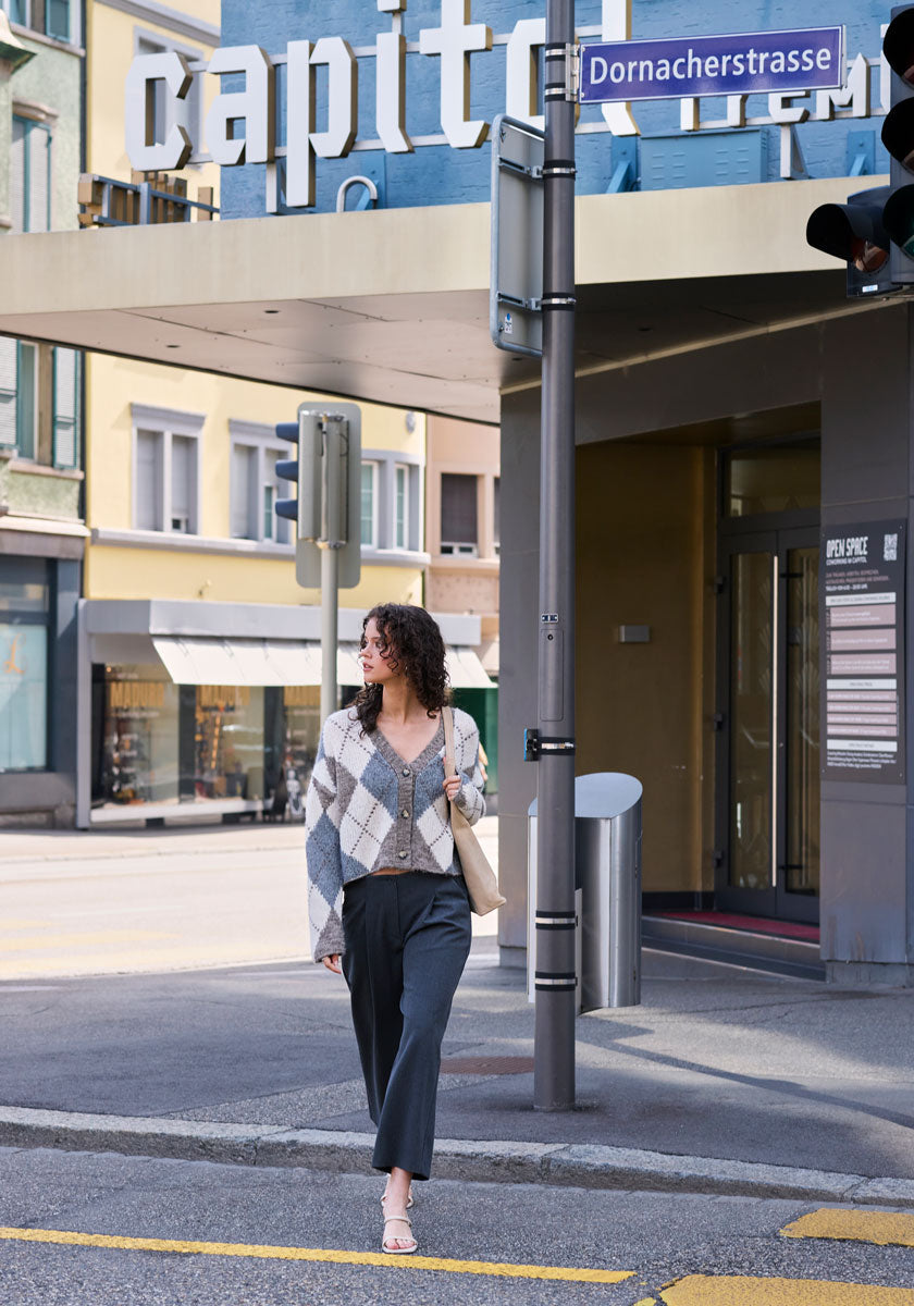 A woman with curly hair and glasses walks on a city sidewalk, wearing gray pants, sandals, and a plaid Princess Goes Hollywood Wolle-Alpaka-Mix Strickjacke. She carries a beige bag. Above her is a blue Dornacherstrasse sign and the capit cinema sign.