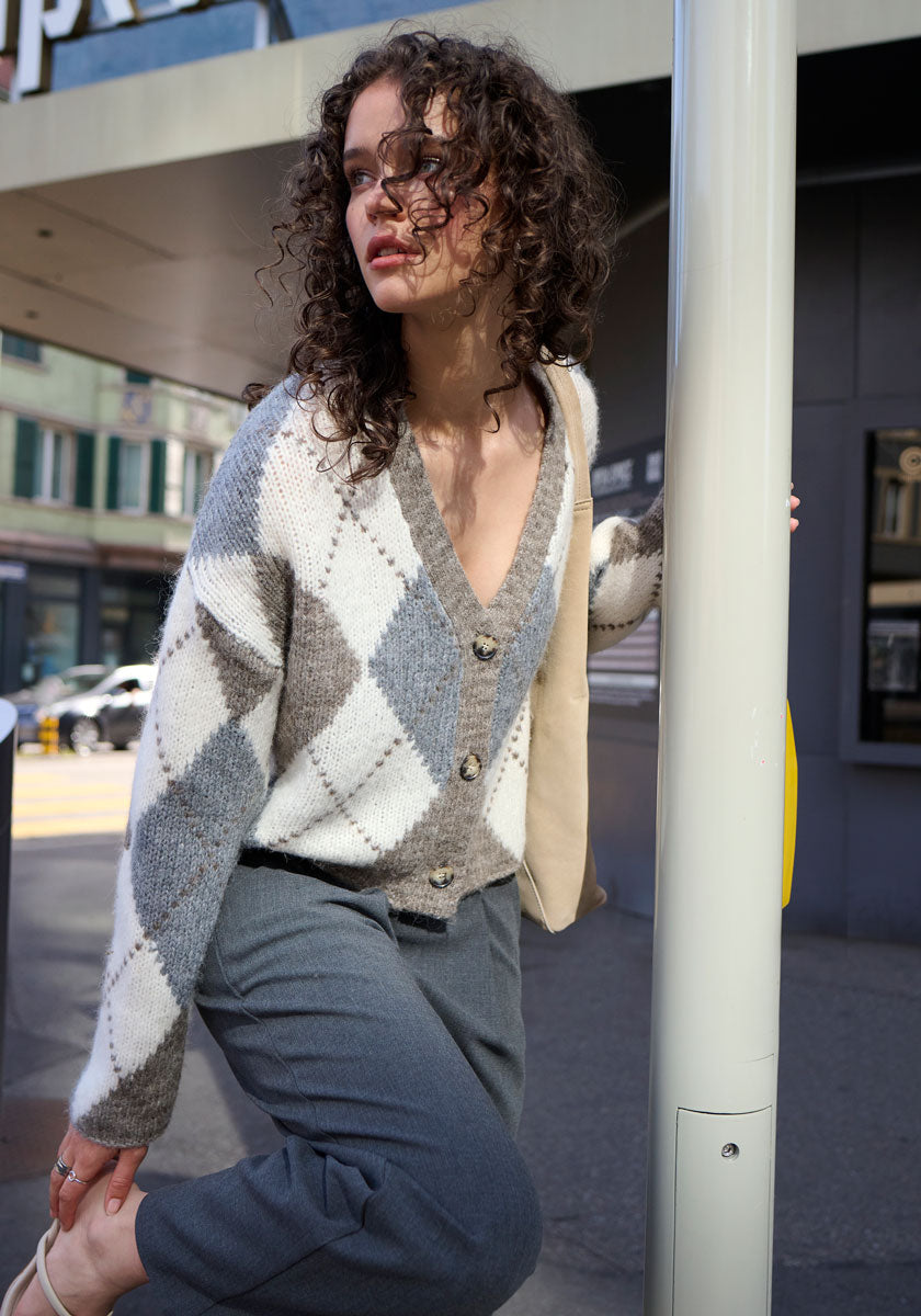 A woman with curly hair, wearing a gray and white Argyle-Muster Strickjacke by Princess Goes Hollywood and gray pants, leans against a pole on a city street. She looks thoughtfully to the side with urban scenery in the background.