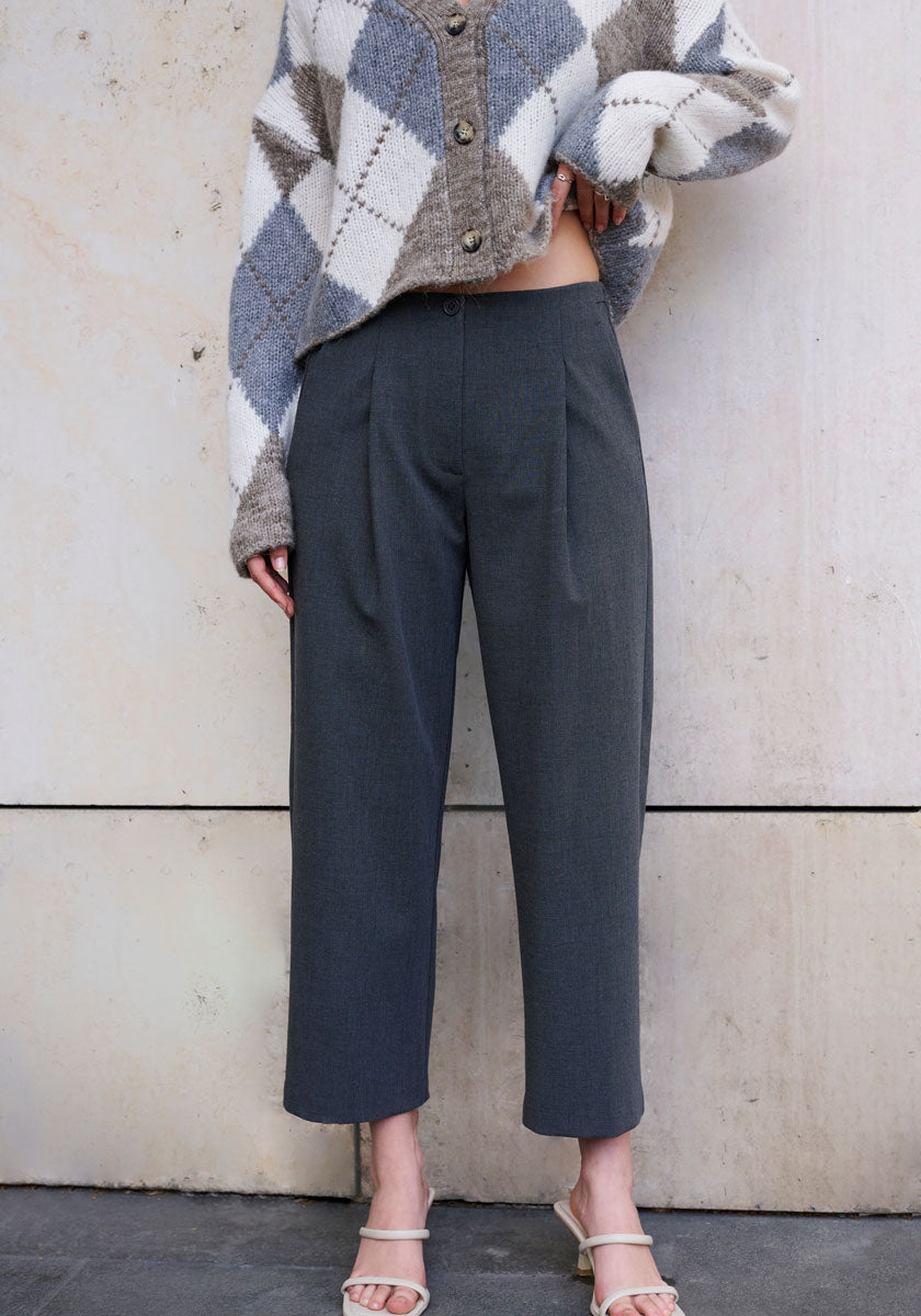 A person stands against a light stone wall wearing a beige and gray argyle sweater by Princess Goes Hollywood and high-waisted, wide-leg, 7/8-length trousers. Their hand rests on their waist; cream open-toe heeled sandals complete the look. Face not visible.