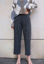 A person stands against a light stone wall wearing a beige and gray argyle sweater by Princess Goes Hollywood and high-waisted, wide-leg, 7/8-length trousers. Their hand rests on their waist; cream open-toe heeled sandals complete the look. Face not visible.