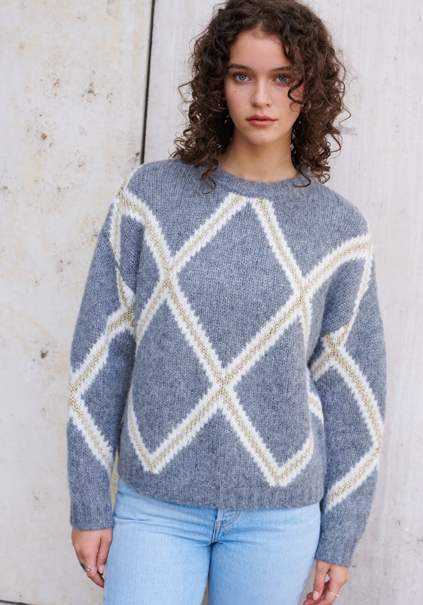 A young woman with curly brown hair stands against a light, textured wall, wearing a Princess Goes Hollywood gray Strickpullover with bold white and beige Argyle-Muster and light blue jeans. She faces the camera directly, arms relaxed at her sides.