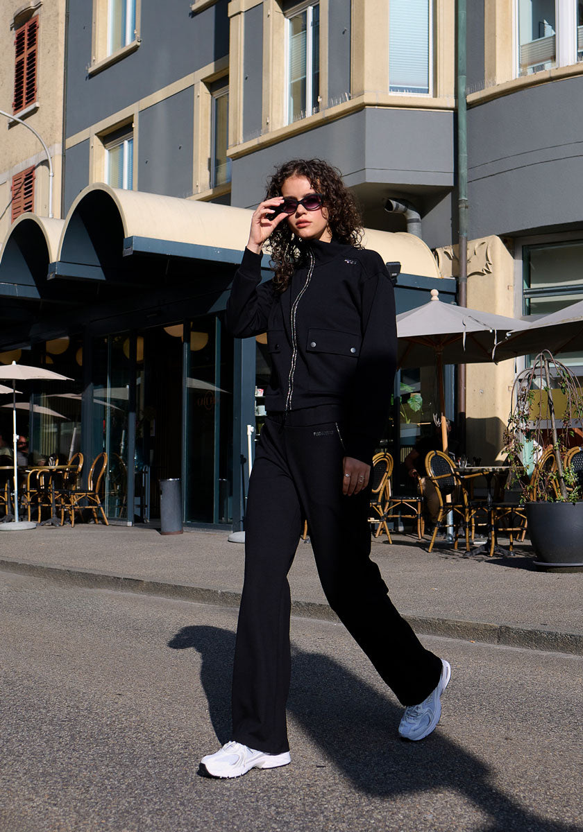 A woman with curly hair walks confidently on a city sidewalk, wearing a black zip-up jacket and Sweathose mit elastischem Bund by Princess Goes Hollywood, white sneakers, and sunglasses. She lifts her sunglasses slightly near a café.