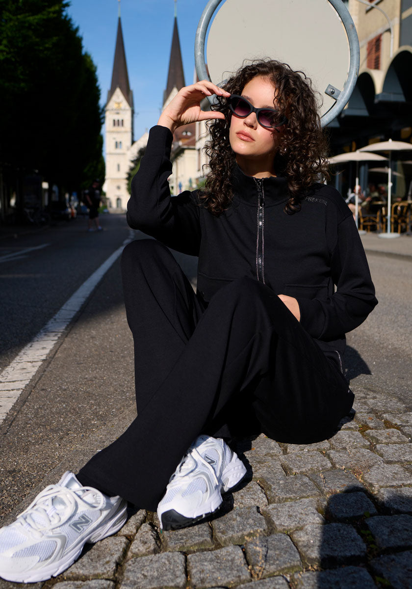 A woman with curly hair wears sunglasses and a black Princess Goes Hollywood zip-up tracksuit with Sweathose featuring elastischer Bund, paired with white sneakers. She sits on a cobblestone sidewalk near a street, sign, and church with two spires.