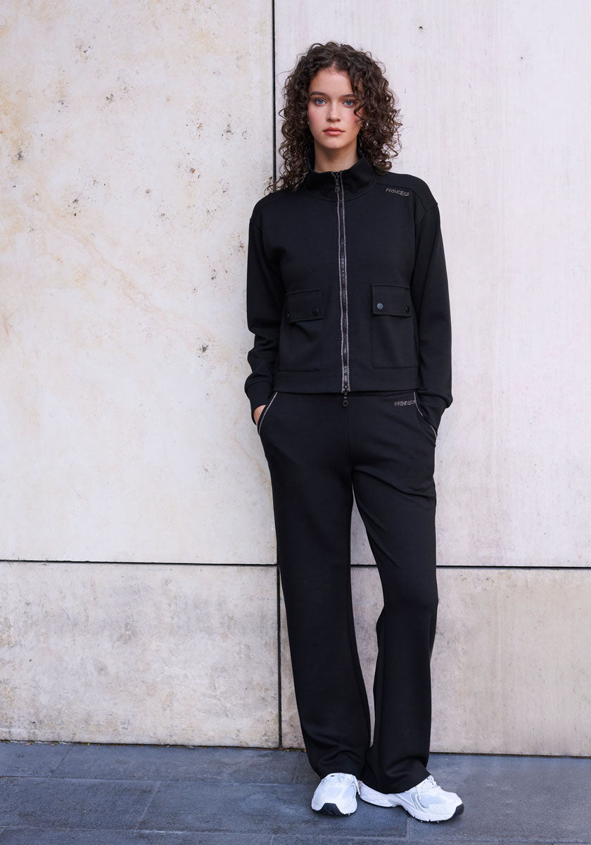 A young woman with curly brown hair stands by a light stone wall, wearing Princess Goes Hollywood’s black zip-up jacket with strass details, matching sweatpants with an elastic waistband, and white sneakers. Her hands are in her pockets as she gazes at the camera.