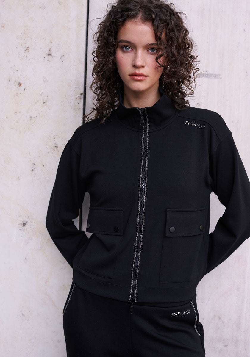 A young woman with curly brown hair stands against a pale, textured wall, dressed in a black Sweatjacke with front pockets and matching pants by Princess Goes Hollywood, her calm expression adding to the modern princess vibe.