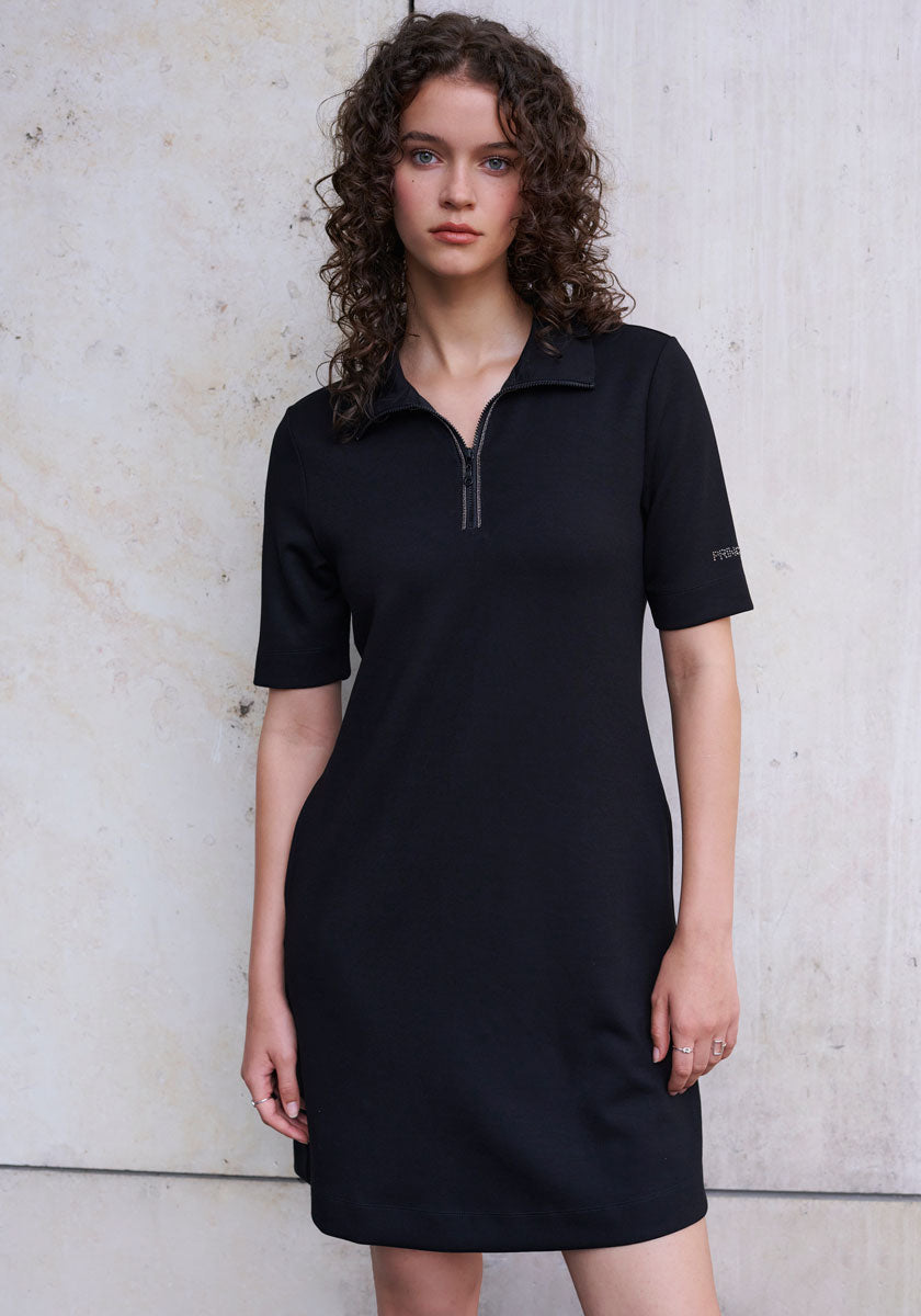 A woman with curly brown hair stands against a marble wall wearing a short-sleeved Mini Sweatkleid with zip-up collar from Princess Goes Hollywood, looking at the camera with a neutral expression and her hands at her sides.