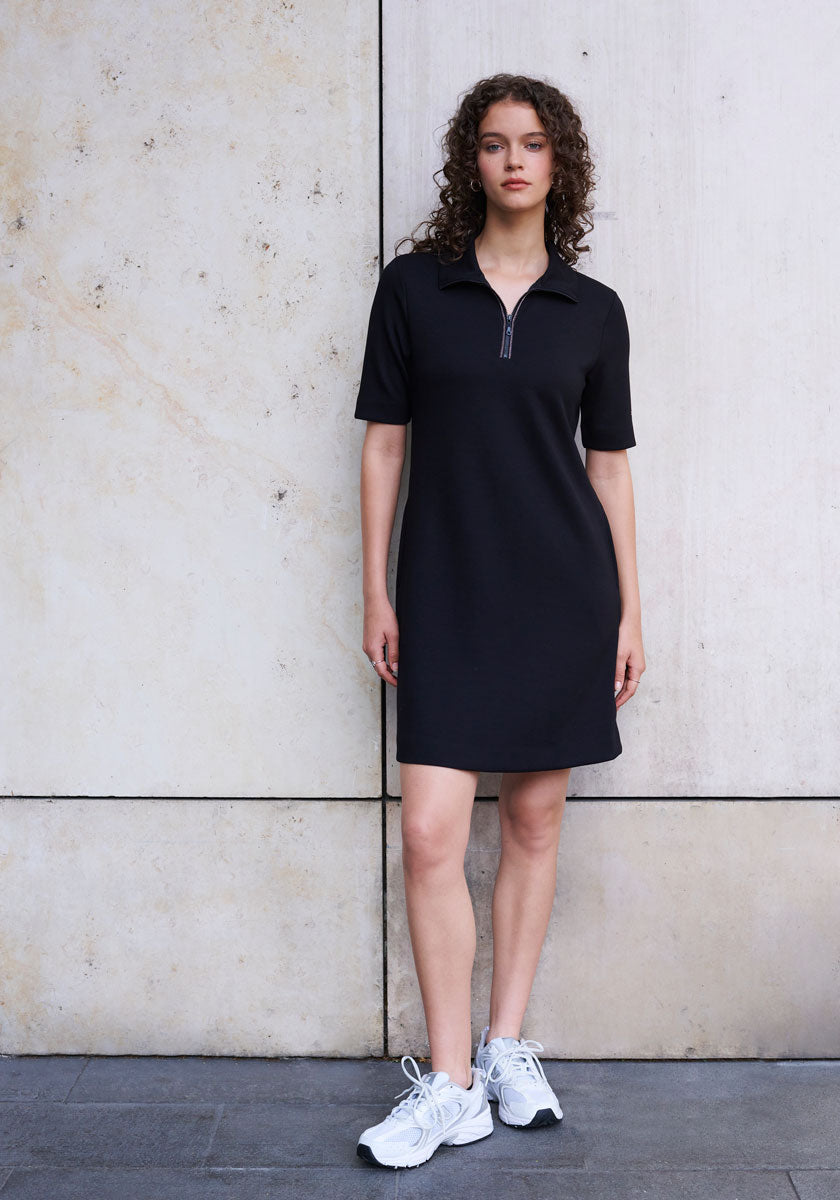 A woman with curly brown hair stands by a beige stone wall, wearing a black Princess Goes Hollywood Mini Sweatkleid with zip collar and white sneakers, arms relaxed by her sides as she faces the camera neutrally.