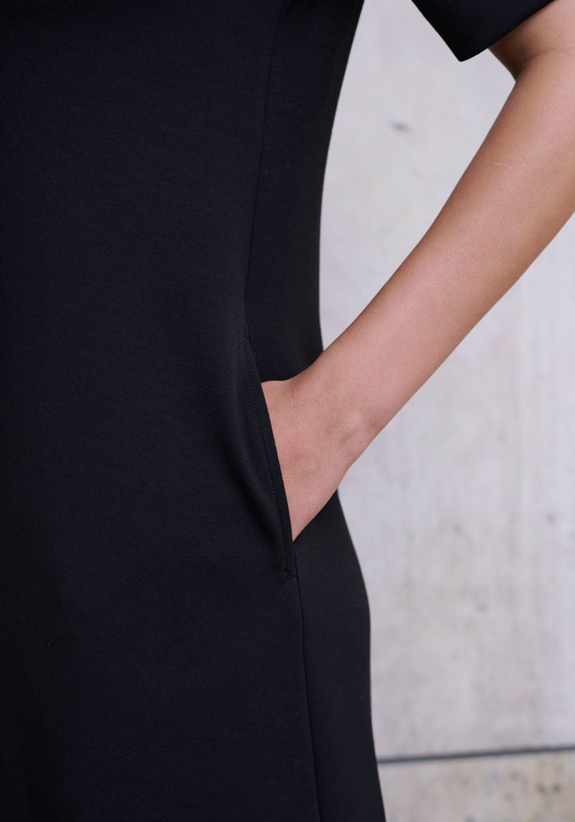 A person models the Princess Goes Hollywood black Mini Sweatkleid against a light, textured wall. Their right hand is in a side pocket, accentuating the dress’s seamless design. The photo shows only the torso and part of one arm.