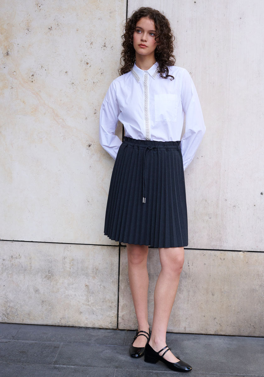 A young woman with curly brown hair stands against a beige wall, wearing a white shirt with trim, a high-waisted schwarzer Plissee Rock mit elastischer Taille from Princess Goes Hollywood, and black Mary Jane shoes. She gazes to the side, arms behind her back.