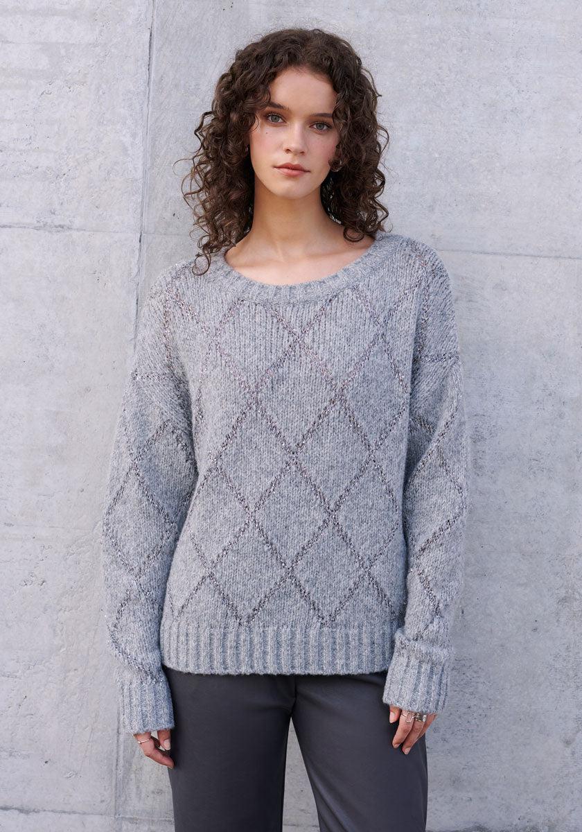 A woman with curly brown hair stands before a light gray concrete wall, wearing a Princess Goes Hollywood Strickpullover mit Argyle-Muster and dark gray pants. She gazes ahead neutrally, hands resting at her sides.