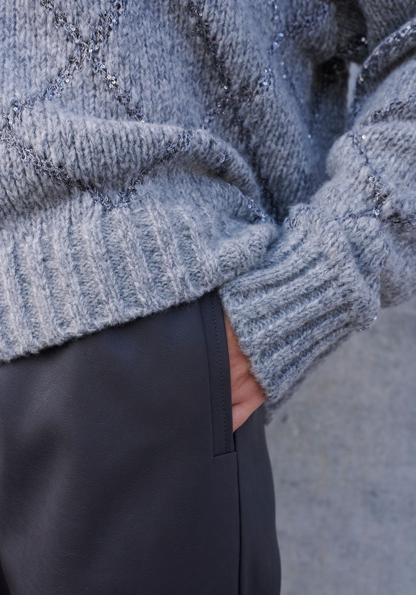 A close-up of a person in a chunky, light gray diamond-patterned sweater and dark gray 7/8-length Kunstlederhose by Princess Goes Hollywood, their right hand tucked in the pocket, set against a soft neutral gray background.