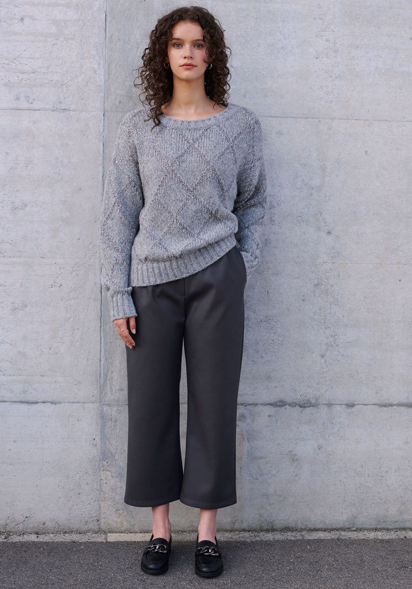 A woman with curly brown hair stands against a gray wall, wearing a textured gray sweater, dark gray 7/8-length Kunstlederhose with elastic waistband from Princess Goes Hollywood, and black loafers. Her hands are in her pockets as she looks at the camera.