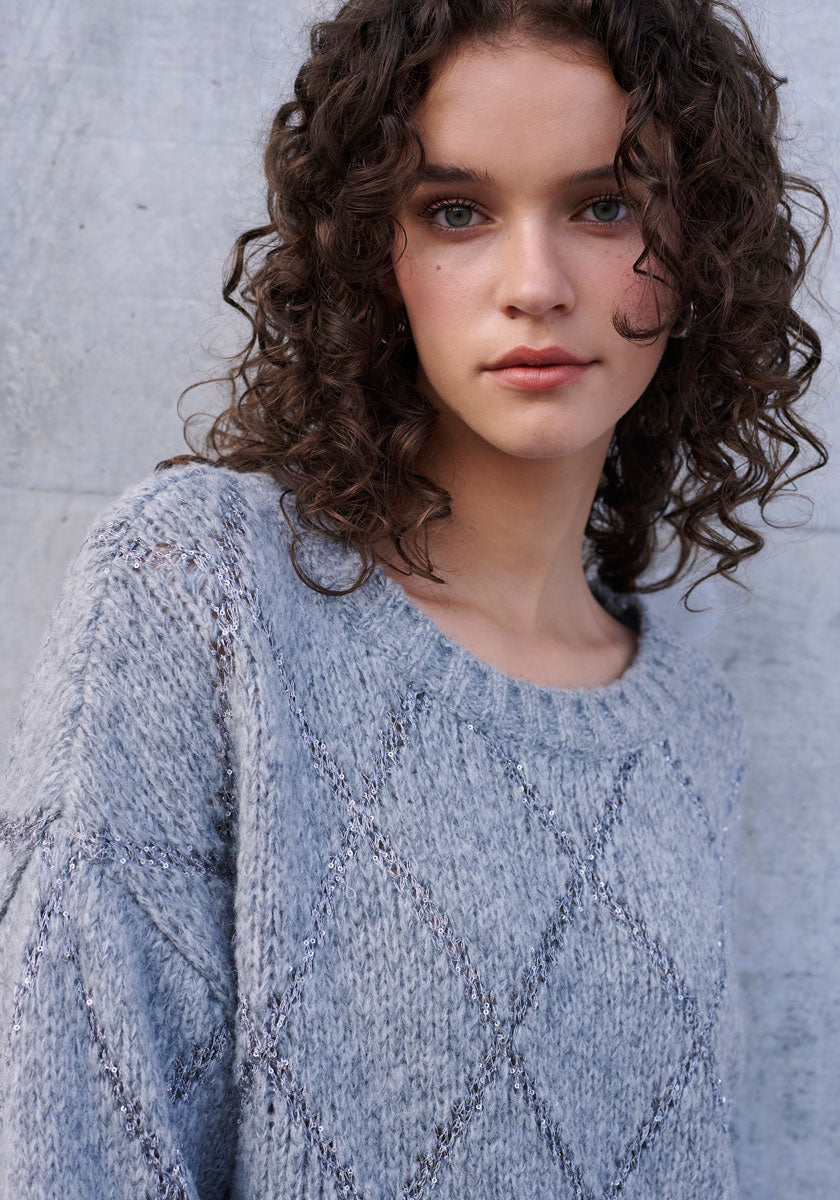 A young woman with curly brown hair and blue eyes stands before a textured gray wall, wearing a light gray Argyle-Muster Strickpullover by Princess Goes Hollywood. She gazes softly at the camera with a neutral expression.
