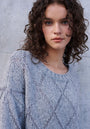 A young woman with curly brown hair and blue eyes stands before a textured gray wall, wearing a light gray Argyle-Muster Strickpullover by Princess Goes Hollywood. She gazes softly at the camera with a neutral expression.