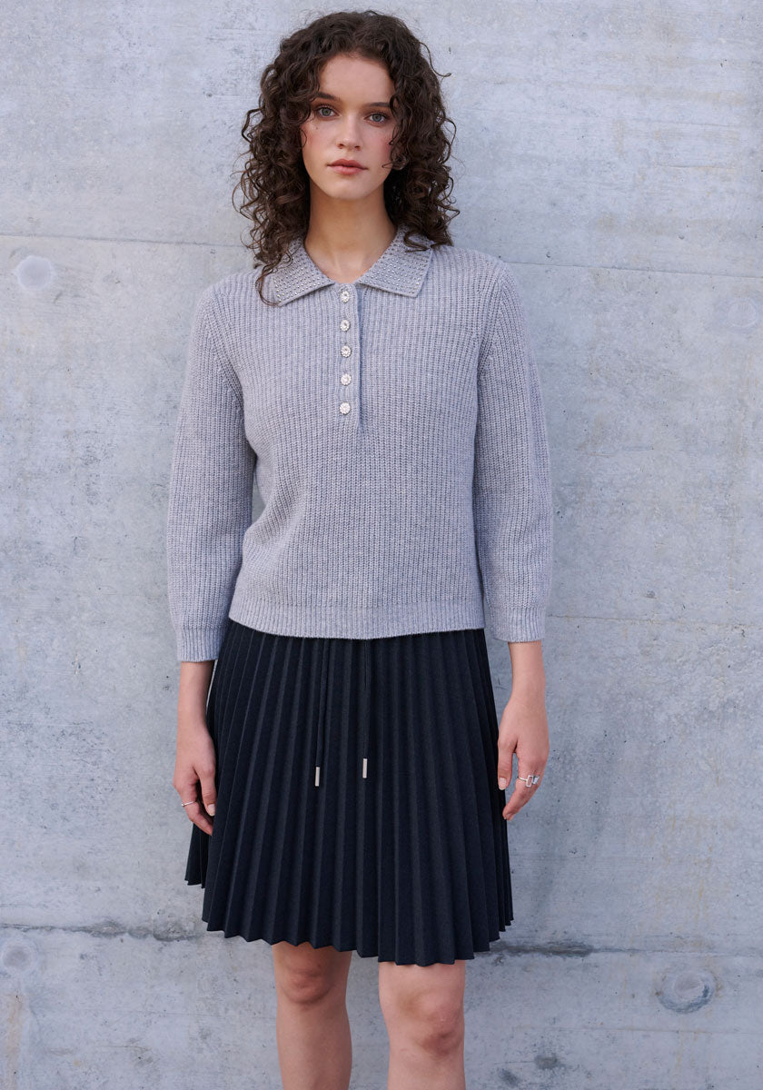 A woman with curly brown hair stands against a light concrete wall, wearing a ribbed, long-sleeve gray Polokragen sweater with button placket from Princess Goes Hollywood and a black pleated skirt. She faces forward, arms at her sides, expression neutral.